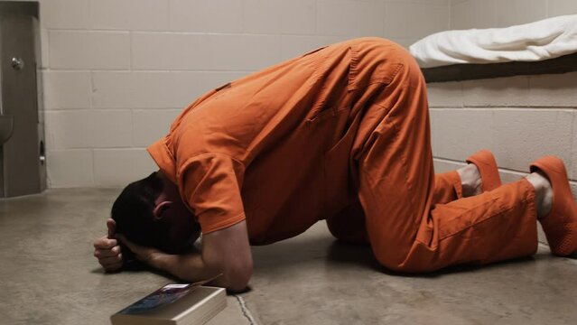 A sad, depressed man in orange prison uniform laying on prison cell floor weeping, crying, praying. Criminal prisoner, inmate in orange jumpsuit sitting in prison cell.