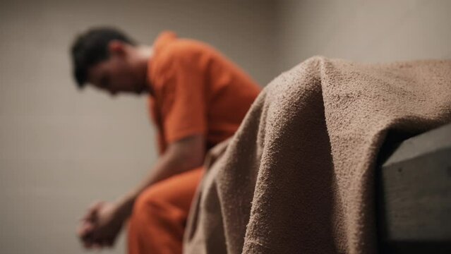 A sad man, convicted criminal, prison inmate in orange uniform sitting on bed. A depressed attractive, handsome man, criminal prisoner, inmate in orange jumpsuit sitting in prison cell.