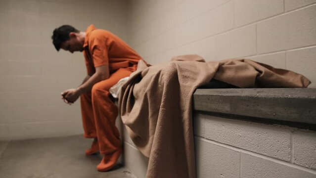 A sad man, depressed attractive, handsome man, criminal prisoner, inmate in orange jumpsuit sitting in prison cell on bed.