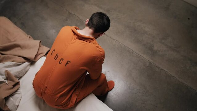 A prison inmate sitting on his bed in prison cell. Sad, depressed attractive, handsome man, criminal prisoner, inmate in orange jumpsuit sitting in prison cell.
