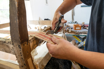 Upholsterer with traditional craft skills shortens a new strap on an old chair with scissors in his workshop, copy space, selected focus