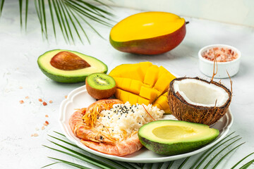 tropical bowl with avocado, prawns, rice, mango, kiwi and coconut, tropical food on the sea beach on light background with tropical leaves. Healthy food, clean eating, Buddha bowl, top view