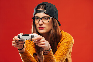 Attractive young hipster in snapback and eyeglasses sitting and holding gaming controller on red...