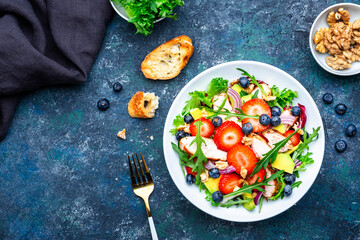 Healthy summer salad with strawberry, grilled chicken slices, fresh herbs with blueberries, avocado and walnuts, blue stone table., top view