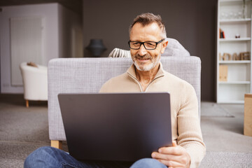 An older man sits relaxed on the floor in the living room and looks at his laptop - 615071833