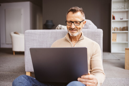 An older man sits relaxed on the floor in the living room and looks at his laptop