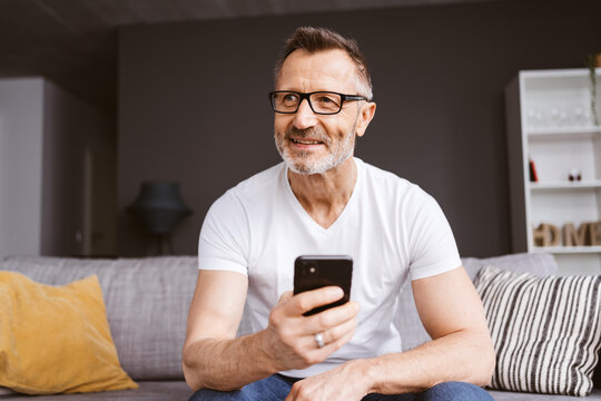 Older Man in White T-Shirt Sitting with Smartphone on the Sofa, Looking to the Side