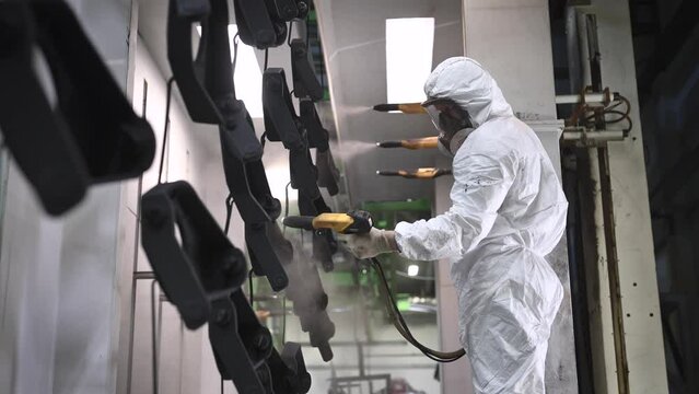 A worker in full protective gear is operating on a production line. Using a special gun, the man is painting parts. This process is an essential part of the manufacturing process