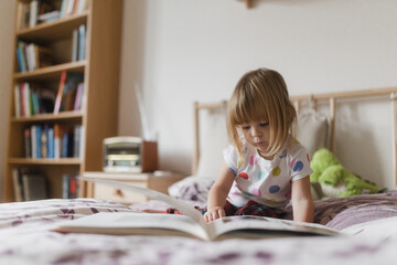 Cute little girl reading book lying on bed.