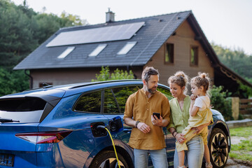 Family with little girl standing in front of their house with solar panels on the roof, having...