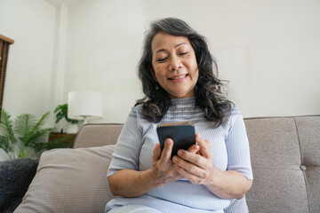 Smiling older asian woman sit rest on couch at home text or message online on modern cellphone gadget. Happy mature female relax browse internet on smartphone device. Elderly technology concept