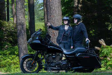 man and women motorcycle riders parked near mountain trees pointing at something in the distance. Black motorcycle and its two riders enjoying the mountain woodlands.