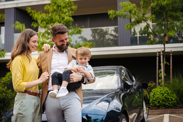 Happy family standing in front their car and charging electric car on the street.