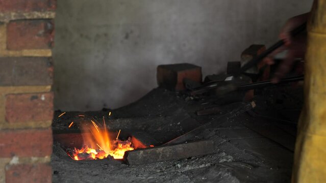 Blacksmith stokes a coal fire and pounds a red hot peice of metal with a hammer