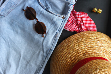 Jeans, gingham top, straw bag and hat, accessories and bowl of strawberries on dark background. Spring or summer picnic outfit. Top view.