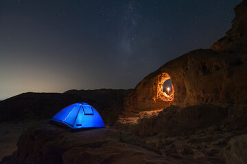 Man standing with a flare light under a stone arch in the desert during the night with the milky way in the background and a tent with light inside on a cliff 