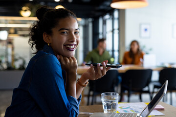 Portrait of happy biracial casual businesswoman using laptop talking on smartphone in office lounge