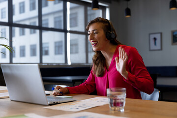 Happy caucasian casual businesswoman making video call using headphones and laptop at desk