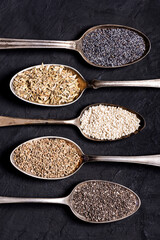 top view of some spoons with variety of organic seeds on black textured background