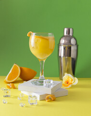 Glass wine glass with an orange cocktail on a white wooden stand. Orange slices, yellow orchid blossom and shaker in the background. Light green background. Ice cubes on a yellow table