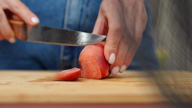 Closeup slicing half of flat peach in slow motion with knife. Female Caucasian hands cutting delicious tasty ripe fruit on cutting board indoors