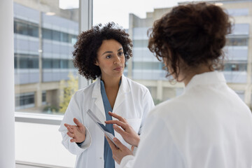 Two female doctors discussing patient treatment in a busy hospital using a digital tablet