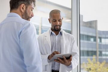 Two male doctors discussing patient treatment in a busy hospital using a digital tablet