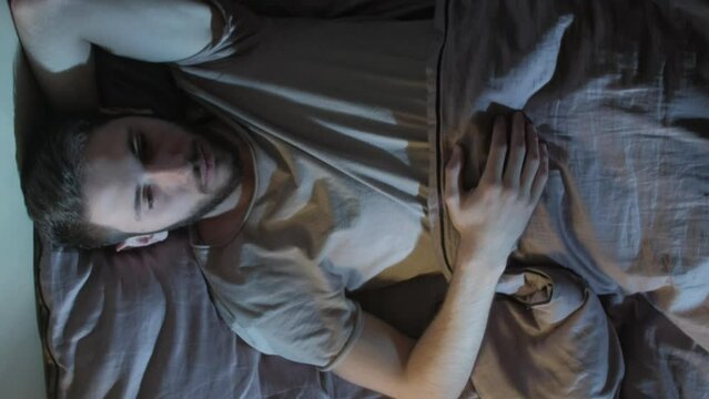 Vertical video. Sleepless night. Insomnia problem. Fatigue disorder. Depressed tired pensive man overthinking awake lying in bed in dark home bedroom.