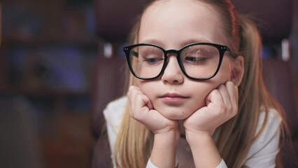 Close up of focused precious child in glasses viewing interesting cartoon on laptop while sitting in soft chair with hands under chin. Concentrated female kid being hypnotized by media.