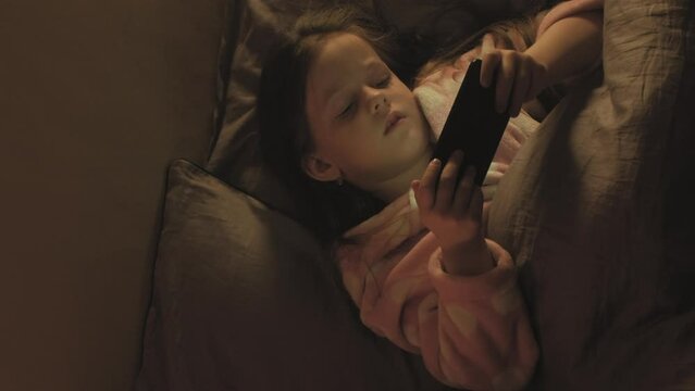 Vertical video. Bedtime movie. Night gadget. Sleepless child. Tired little girl watching cartoons on phone in bed late in dark home bedroom.