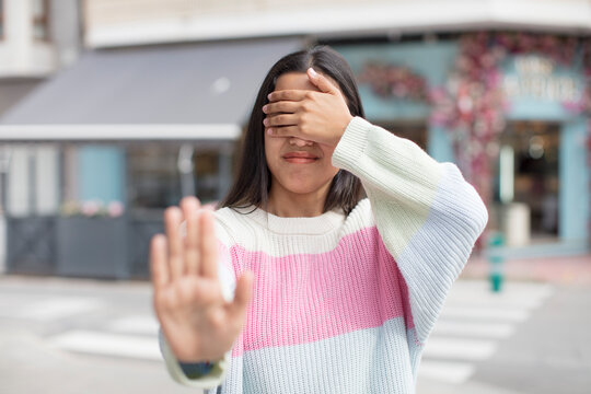 pretty hispanic woman covering face with hand and putting other hand up front to stop camera, refusing photos or pictures