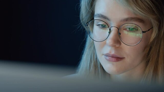 Panning right close up of cheerful woman sitting in front of computer at night and choosing something to watch wearing glasses with screen reflection in them