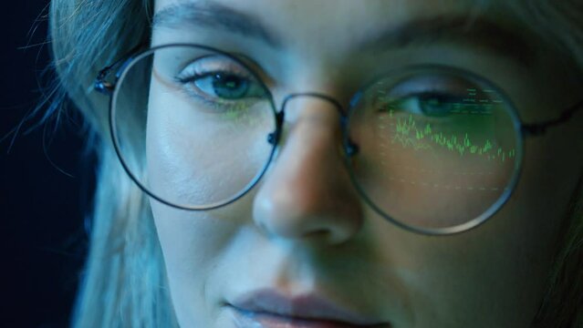 Panning left extreme close up of concentrated young woman sitting in front of computer at night looking at screen studying crypto analytics reflecting in glasses