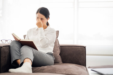 Lady suffering eyestrain reading book sitting on a couch in living room at home