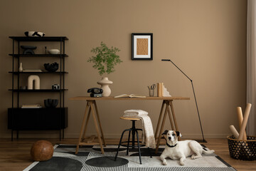 Warm and cozy workplace interior with mock up poster frame, dog, wooden desk, stool, patterned rug, books, pencils, phone, black rack, vase with leaves and personal accessories. Home decor. Template.