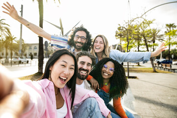 Multi ethnic group of millennial friends taking a selfie in the street. Cheerful multiracial group of young hipsters laughing and taking a picture outside and looking at camera. - obrazy, fototapety, plakaty