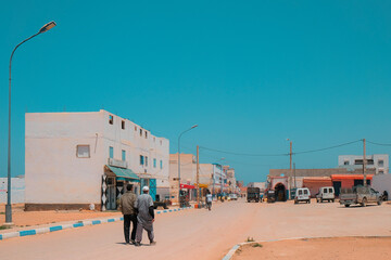 Mirleft, Morocco - two men walk on a dirt road towards a public market. Low buildings and...