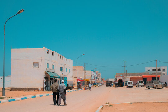 Mirleft, Morocco - two men walk on a dirt road towards a public market. Low buildings and electrical posts on both sides. Vehicles parked outside stores. Local scene in Africa.