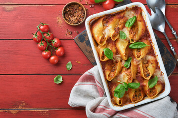 Cannelloni or conchiglioni. Baked stuffed pasta shells with bolognese meat sauce, tomatoes, basil on rustic red wooden table. Traditional Italian bolognese baked pasta with parmesan. Italian cuisine.