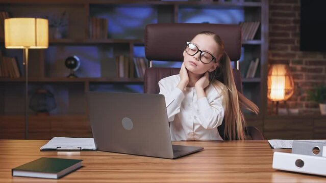 Elegant small child having body ache after working too many hours on laptop. Tired little girl taking short break, massaging neck and making stretching exercises in spacious modern office.