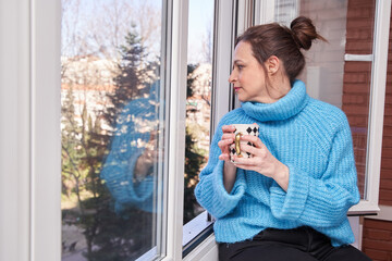 Morning routine. Portrait of happy charming young female enjoying street view through window, drinking good coffee, sitting on windowsill and smiling. Beautiful daydreamer