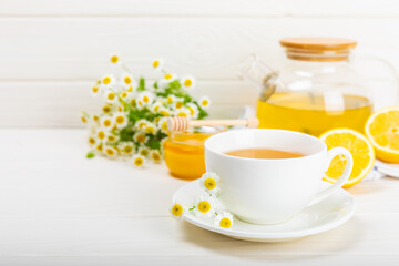 Herbal chamomile tea in a white cup on a white wooden table with honey, lemon and chamomile bouquet. Close-up. Copy space. Useful herbal drinks, immunity tea. Natural healer concept.