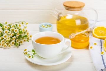 Herbal chamomile tea in a white cup on a white wooden table with honey, lemon and chamomile bouquet. Close-up. Copy space. Useful herbal drinks, immunity tea. Natural healer concept.