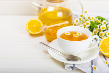 Herbal chamomile tea in a white cup on a white wooden table with honey, lemon and chamomile bouquet. Close-up. Copy space. Useful herbal drinks, immunity tea. Natural healer concept.