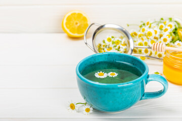 Chamomile herbal tea in a blue cup on a white wooden table with honey, lemon and chamomile bouquet. Close-up. Copy space. Useful herbal drinks, immunity tea. Natural healer concept.Place for text.