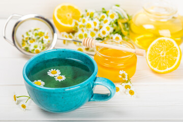 Chamomile herbal tea in a blue cup on a white wooden table with honey, lemon and chamomile bouquet. Close-up. Copy space. Useful herbal drinks, immunity tea. Natural healer concept.Place for text.