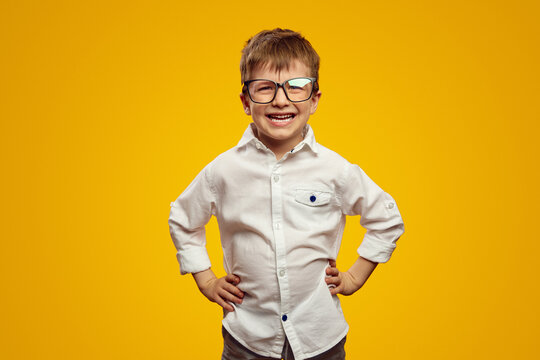 Smart little kid boy wearing nerdy glasses and white shirt smiling for camera while keeping hands on waist against yellow background
