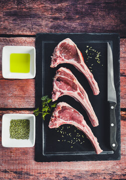 Tres chuletillas de cordero sobre tabla de pizarra con un cuchillo a la derecha y recipientes con aceite y romero a la izquierda