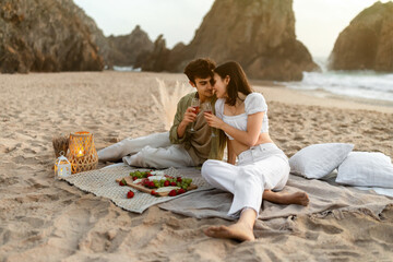 Coastal charm. Lovely couple having picnic on sandy beach at coastline, drinking wine enjoying...