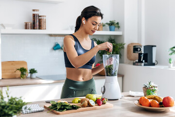 Athletic woman preparing smothie with vegetables and fruits while listening music with earphones in...